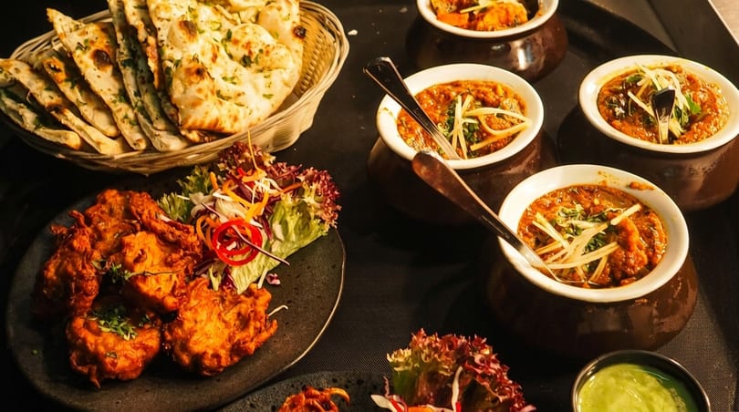 A spread of Indian dishes including naan bread, pakoras, and several bowls of curry on a dark tray.