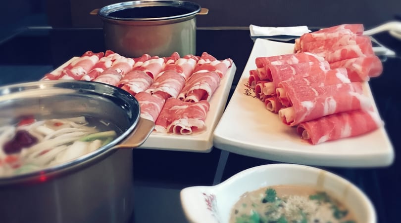 Hot pot meal with two pots, plates of thinly sliced raw meat, and a dipping sauce.
