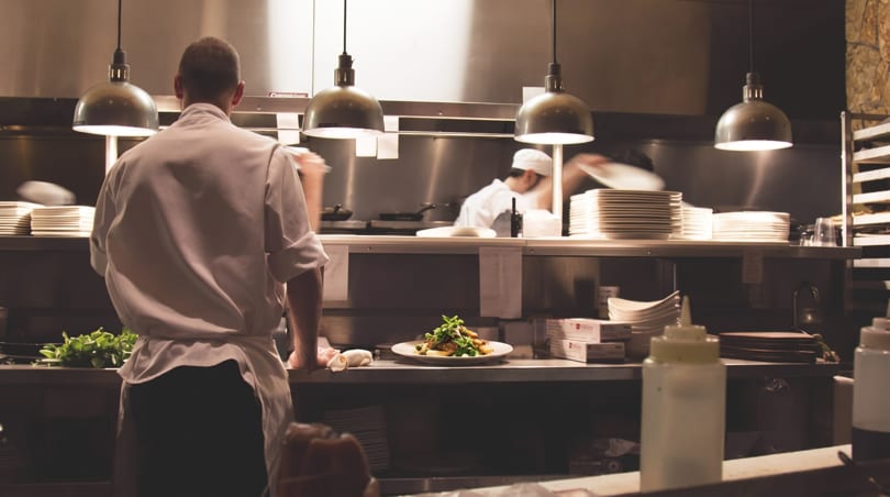 A chef overlooks a plated dish in a professional, bustling restaurant kitchen.