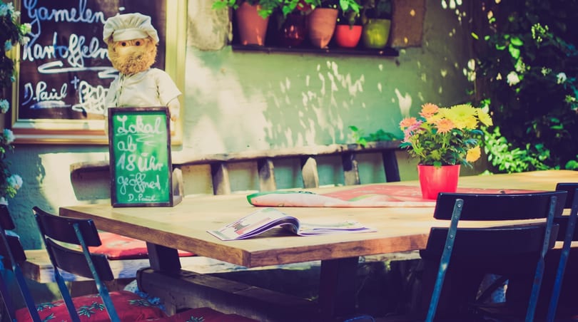 Sunny outdoor restaurant patio with a wooden table, chairs, flowers, and a chef doll by a chalkboard.